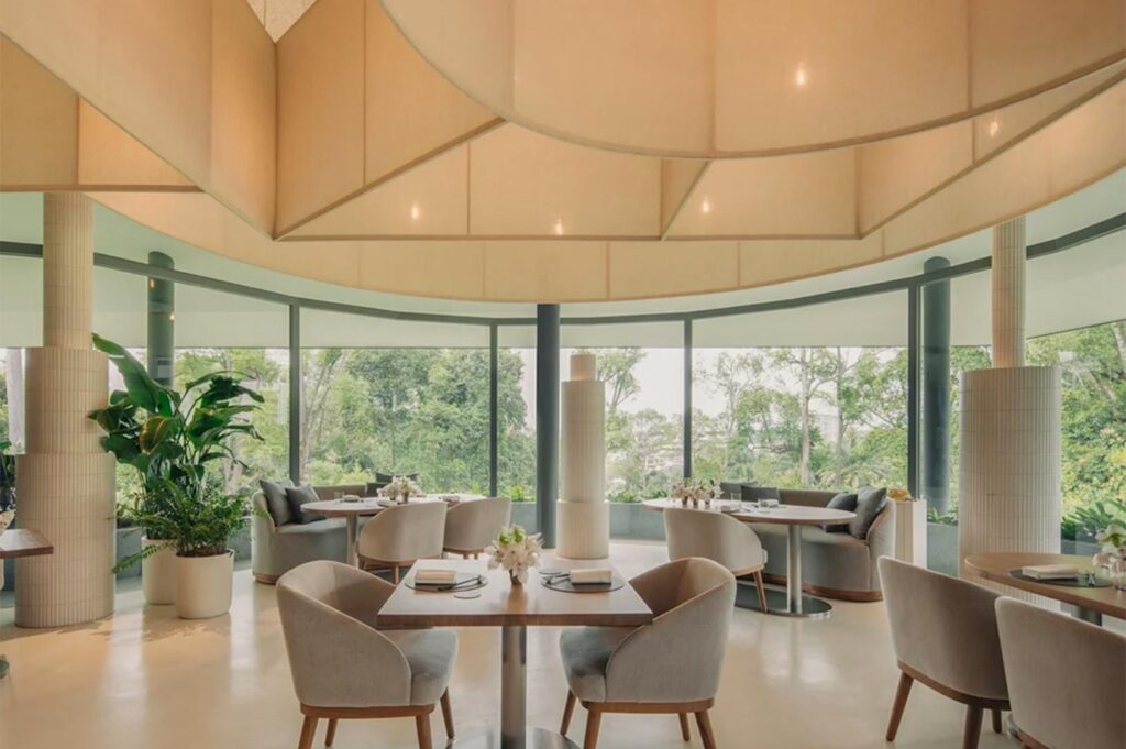 Interior of a minimalist fine dining room featuring floor-to-ceiling curved windows, neutral-toned furniture, and a unique geometric fabric ceiling installation.