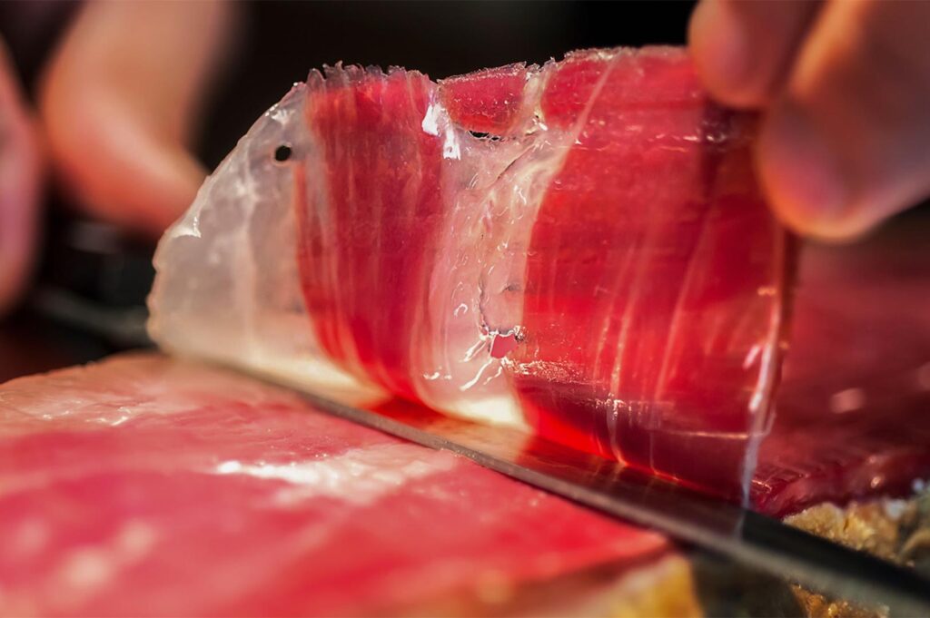 A close-up shot of a professional chef hand-slicing a translucent, marbled strip of Spanish Jamón Ibérico with a long, thin carving knife.
