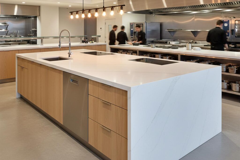 A professional chef's kitchen showcasing a large white marble waterfall island with a sink and induction hob, light wood base cabinets, and stainless steel appliances in the background.