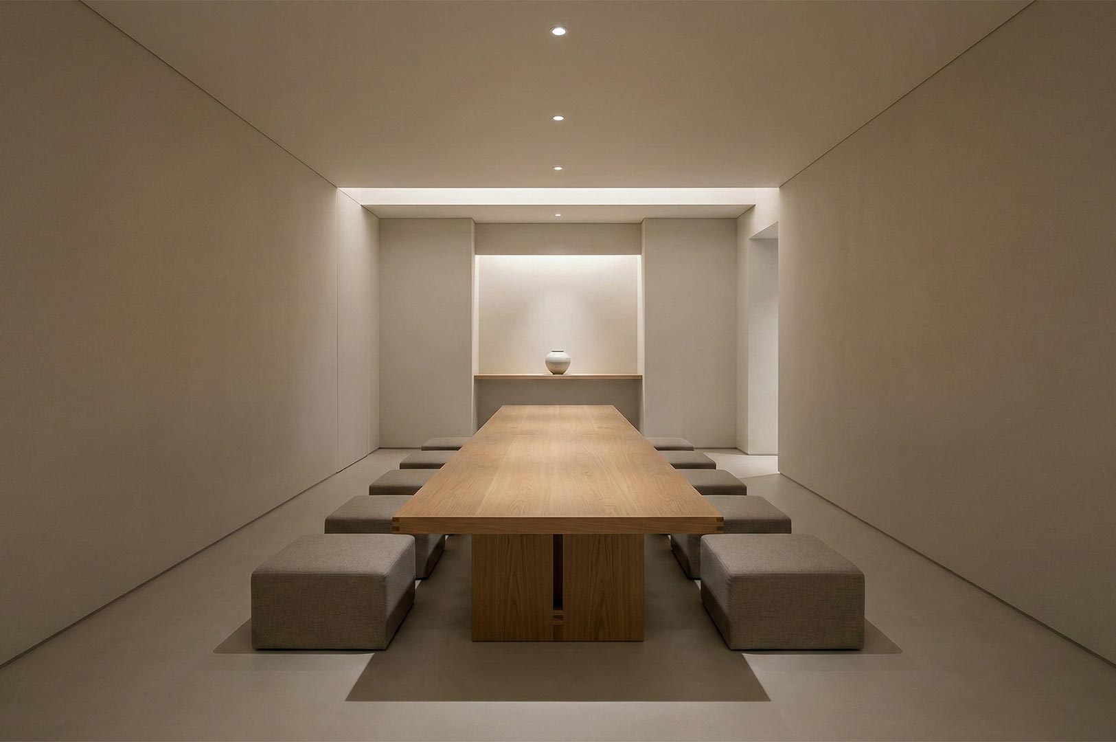 A long light wood tea table with square upholstered stools in a minimalist, beige-toned modern tea room with recessed lighting.