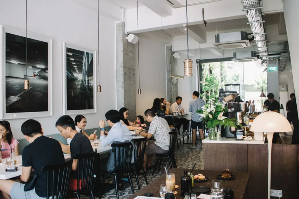 A crowded urban cafe with minimalist white walls, hanging pendant lights, and customers dining at long tables.