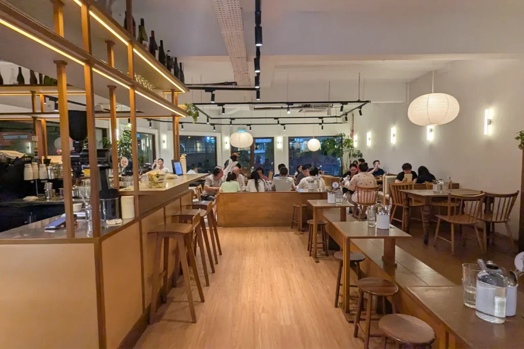 A warm-lit modern cafe interior featuring wooden bar stools, minimalist furniture, paper lantern pendant lights, and customers seated at tables.
