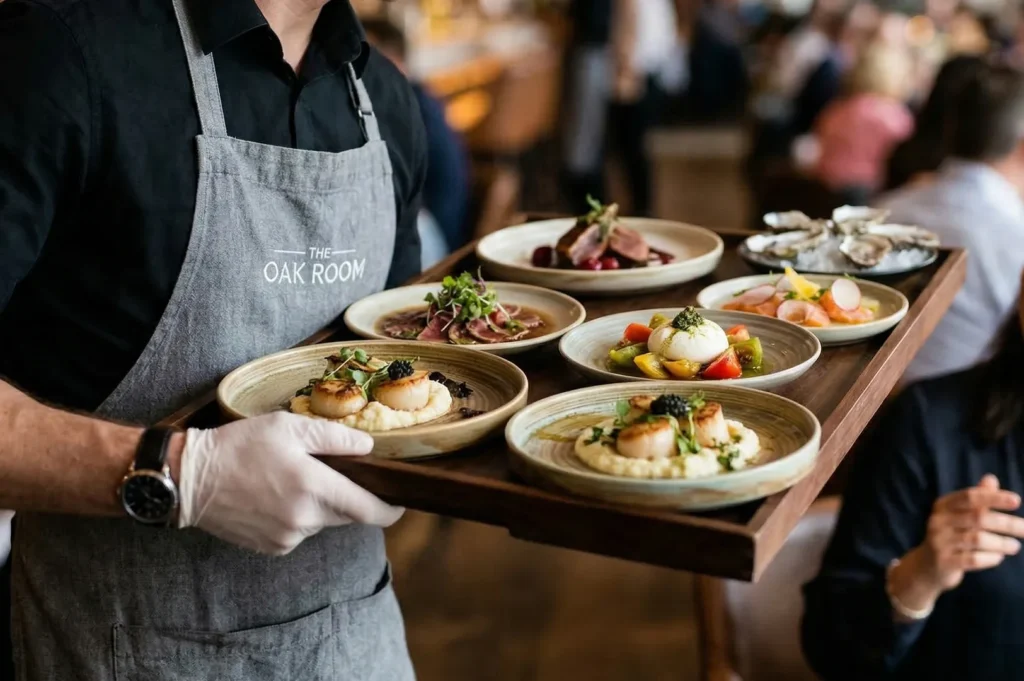 A close-up of a server at The Oak Room carrying a wooden tray with gourmet dishes, including seared scallops and burrata salad.