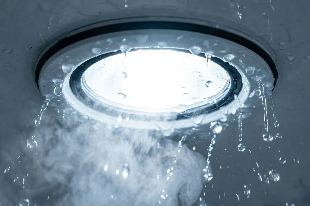 A waterproof recessed LED light fixture being sprayed with water and steam, demonstrating IP-rated durability.