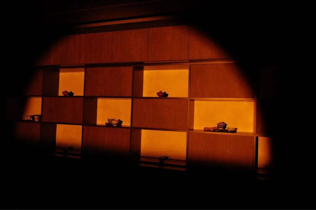 A wide shot of a luxury dark wood display cabinet with glowing amber backlit recessed shelves showcasing delicate tea ware.