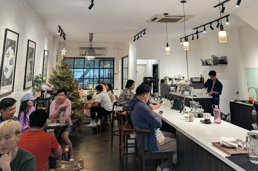 A bustling specialty coffee bar with customers sitting at a long white counter and a barista preparing drinks.