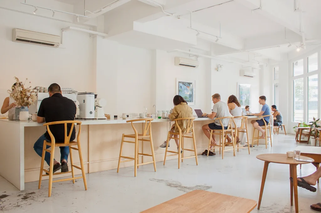 A bright, white-walled minimalist cafe with patrons working on laptops at a long wooden bar.