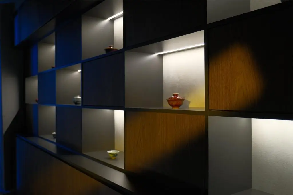 A side angle view of a contemporary shelving unit with spotlighting on individual ceramic tea bowls and jars against a dark wood finish.