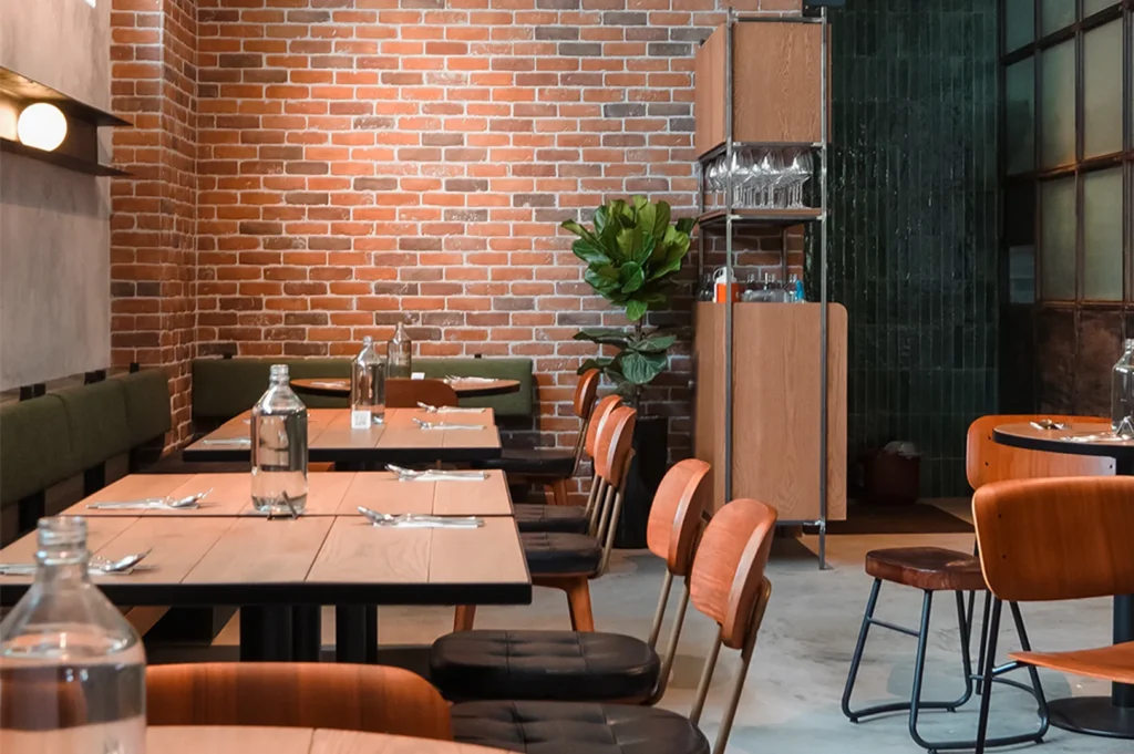 A cozy restaurant interior with an exposed red brick wall, wooden tables, and green banquette seating.