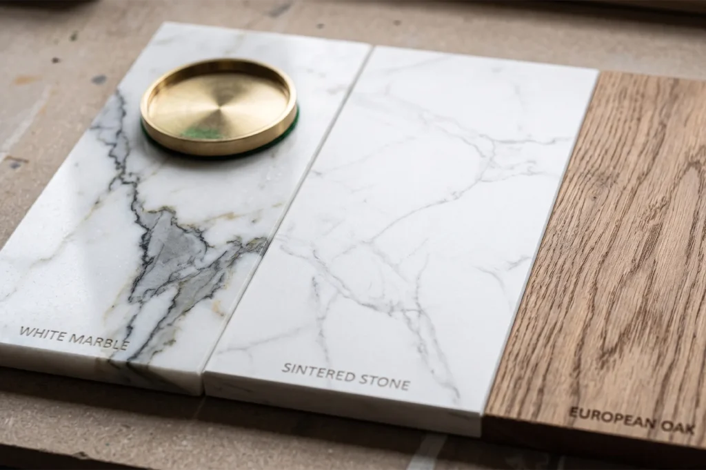 Close-up of interior design material swatches featuring White Marble, Sintered Stone, and European Oak wood with a brass accent.