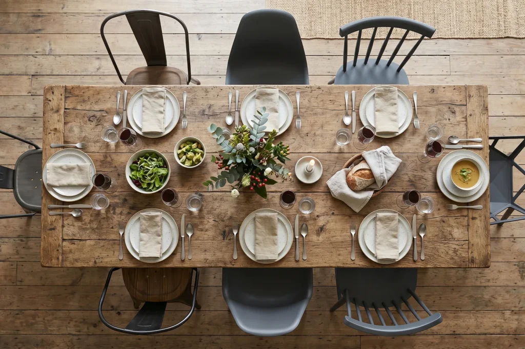 Top-down view of a large rustic farmhouse dining table set for eight people with neutral linens, green salad, wine, and a variety of mismatched grey and metal chairs.
