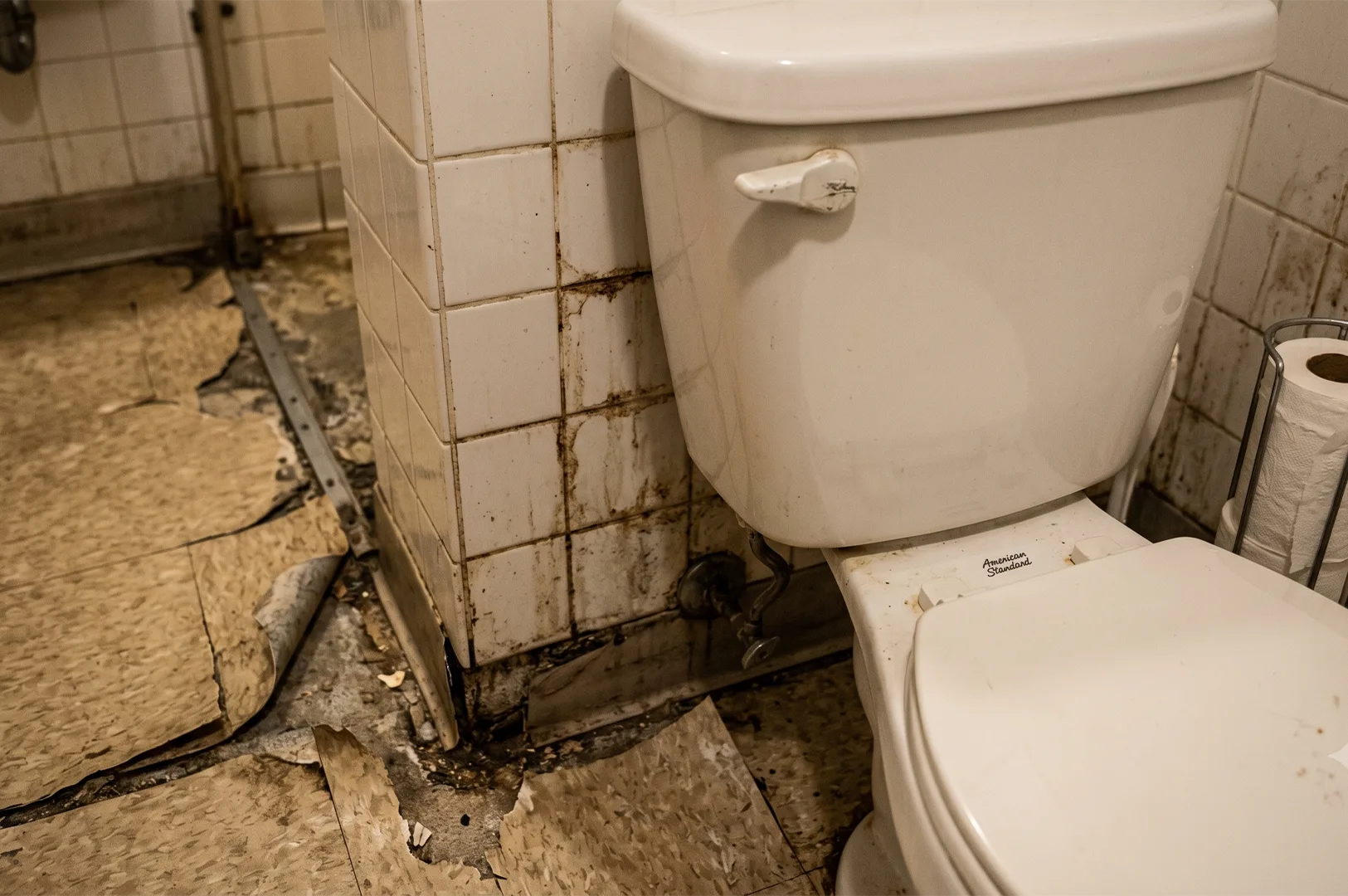 A severely dilapidated bathroom before renovation, showing peeling vinyl floor tiles, water damage stains on white wall tiles, and an old rusted toilet base.