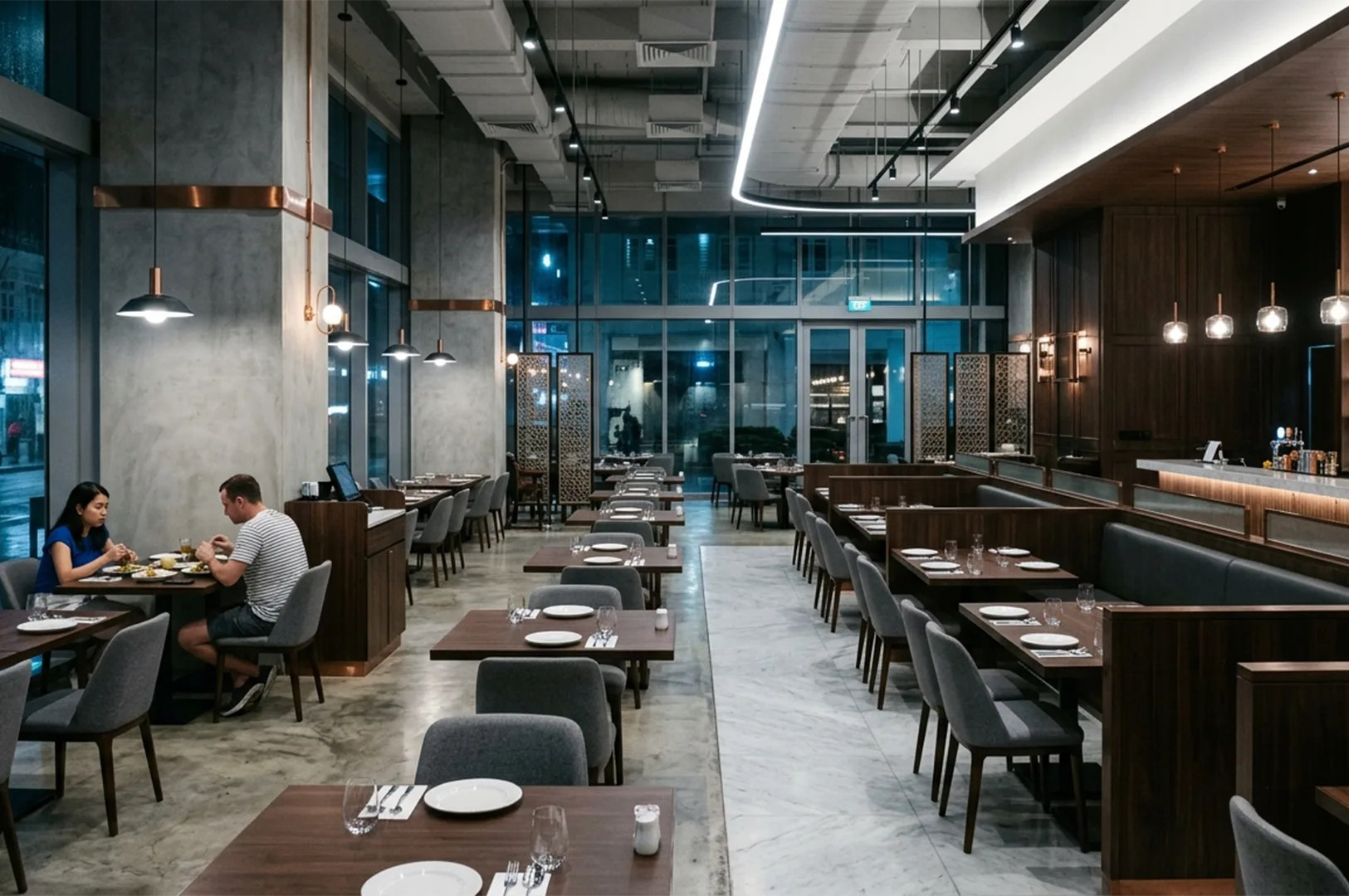 Spacious modern industrial restaurant interior featuring high ceilings, concrete pillars, sleek wooden tables, grey chairs, and ambient pendant lighting with a couple dining in the background.