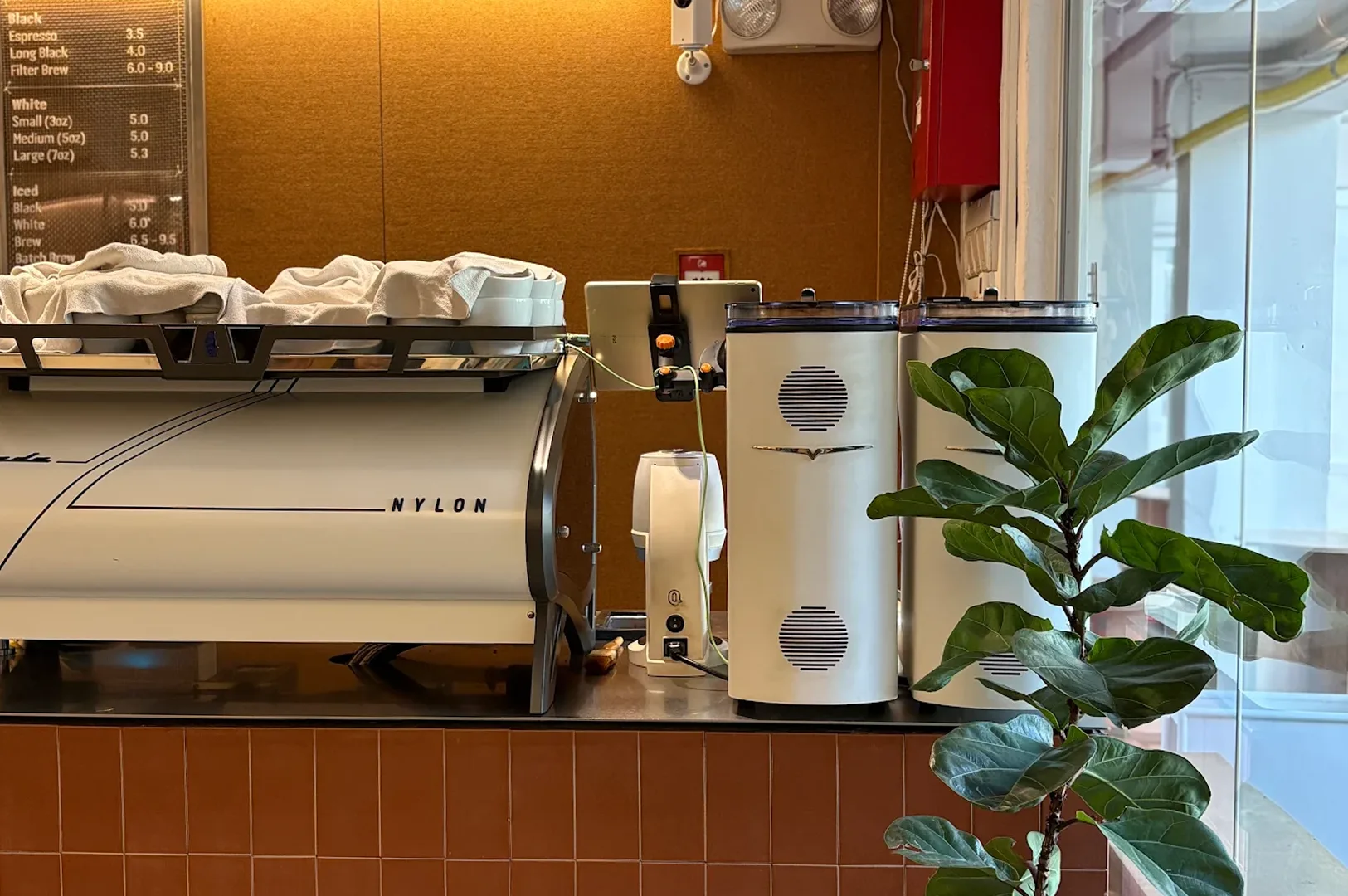 Professional white Nylon branded espresso machine and commercial coffee grinders on a brown tiled counter next to a fiddle-leaf fig plant.