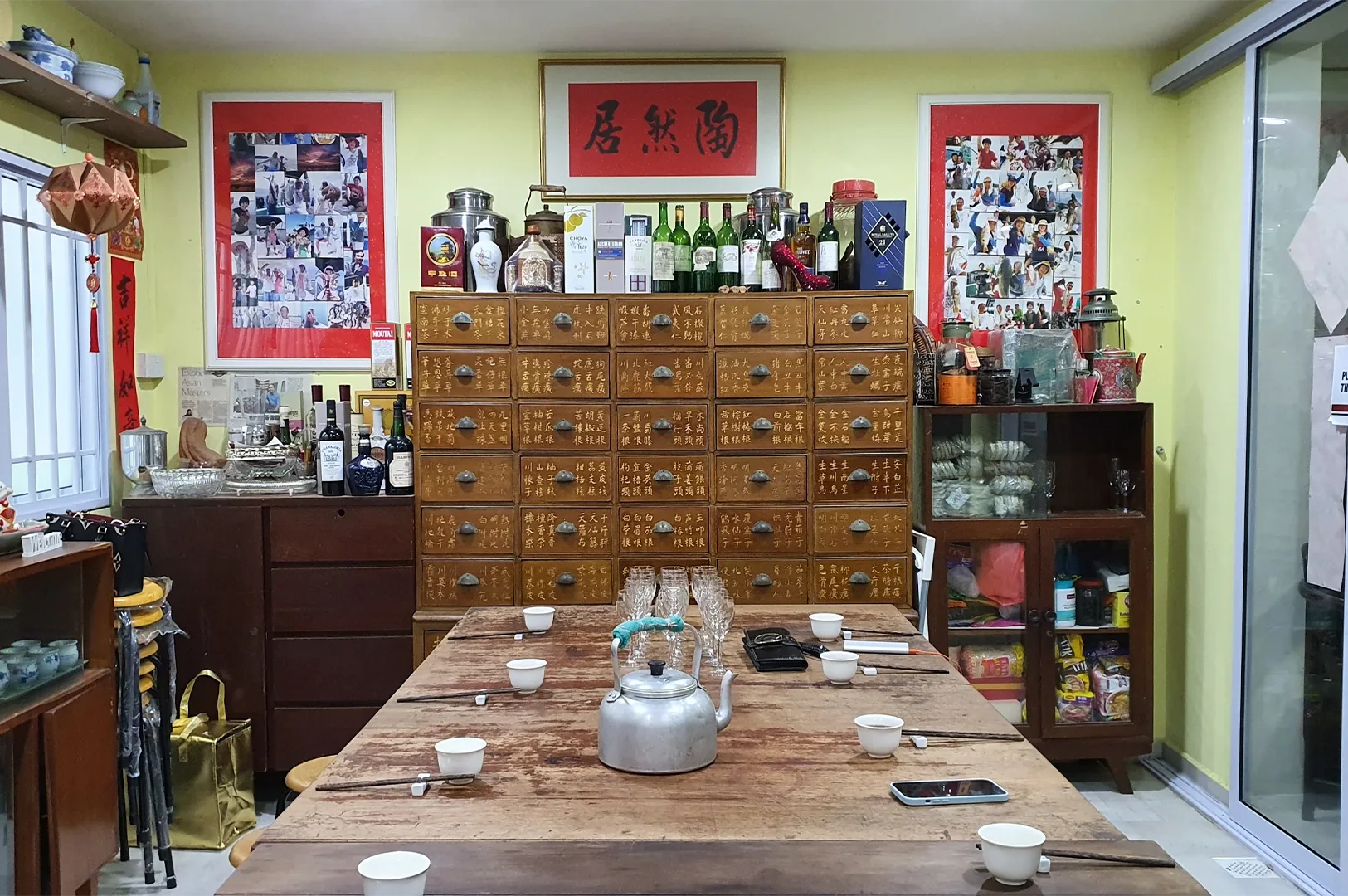 An antique wooden Chinese herbal medicine cabinet with labeled drawers serves as a backdrop for a large rustic dining table set with teacups and a metal kettle.