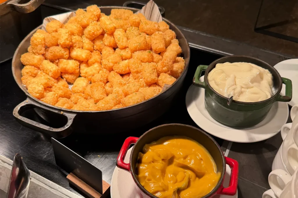 A large bowl of golden brown crispy tater tots served with two pots of creamy cheese dipping sauces at a buffet.