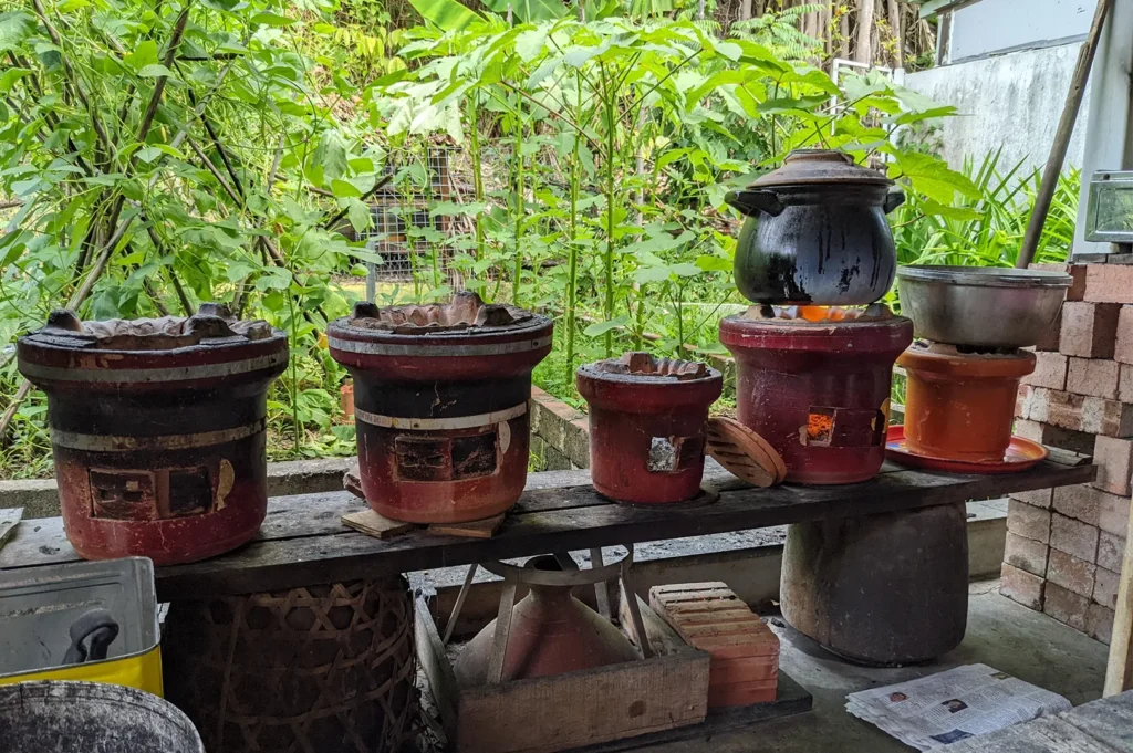 A row of traditional red clay charcoal cooking stoves set up outdoors on a wooden bench, with one pot simmering over an active fire against a lush green background.
