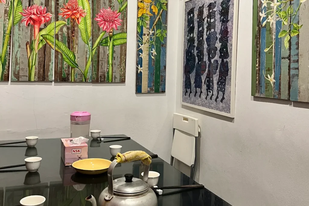 A modern dining table arrangement featuring local Peranakan style floral and figurative paintings on the wall, complete with small teacups and a traditional metal kettle.