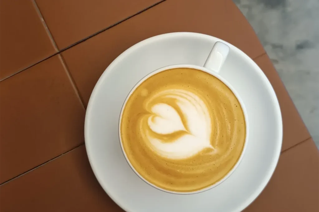 Top-down view of a cup of caffe latte with a heart-shaped latte art design served on a white ceramic saucer over a brown tiled table.