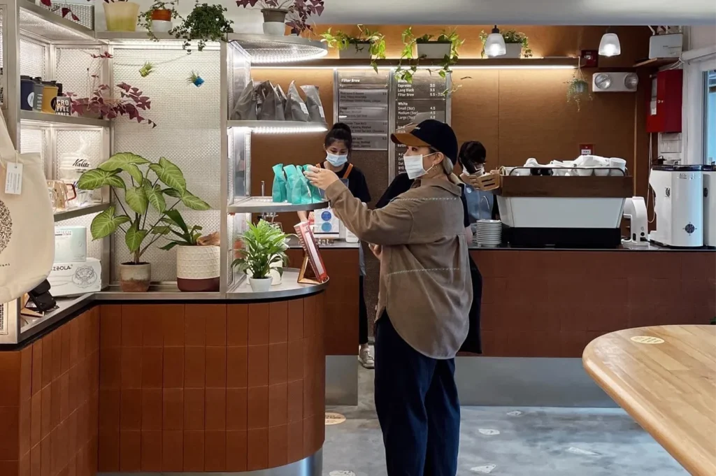 Interior view of Nylon Coffee Roasters cafe with masked baristas working behind a modern brown tiled counter, industrial shelving, and a wooden table.