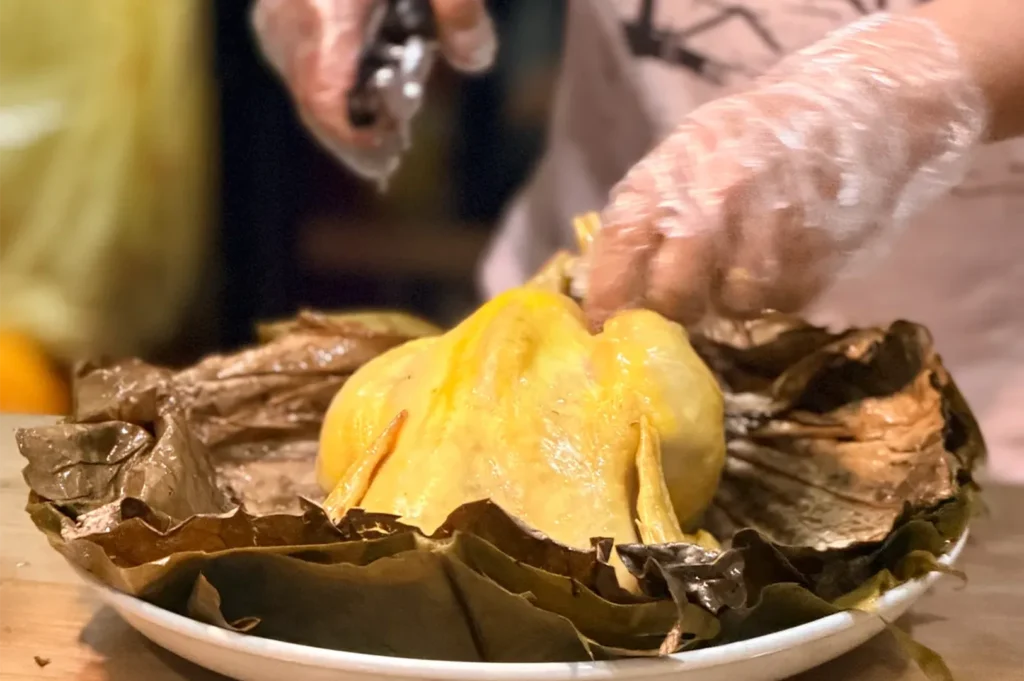 A chef wearing plastic gloves carefully unwrapping or carving a whole tender, golden-yellow steamed Emperor chicken served on a bed of aromatic lotus leaves.