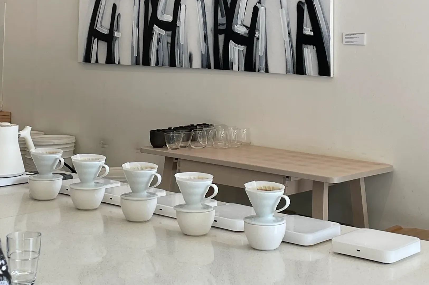 A minimalist pour-over coffee brewing station at Apartment Coffee in Singapore featuring white ceramic V60 drippers on digital scales along a clean white counter under contemporary abstract artwork.