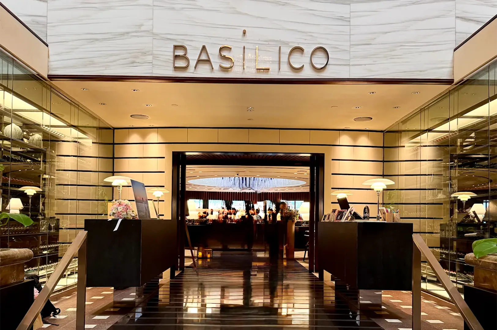 Elegant marble entrance of Basilico Italian restaurant featuring symmetrical dark wooden reception desks, illuminated modern table lamps, and a glimpse of the luxurious dining room interior.