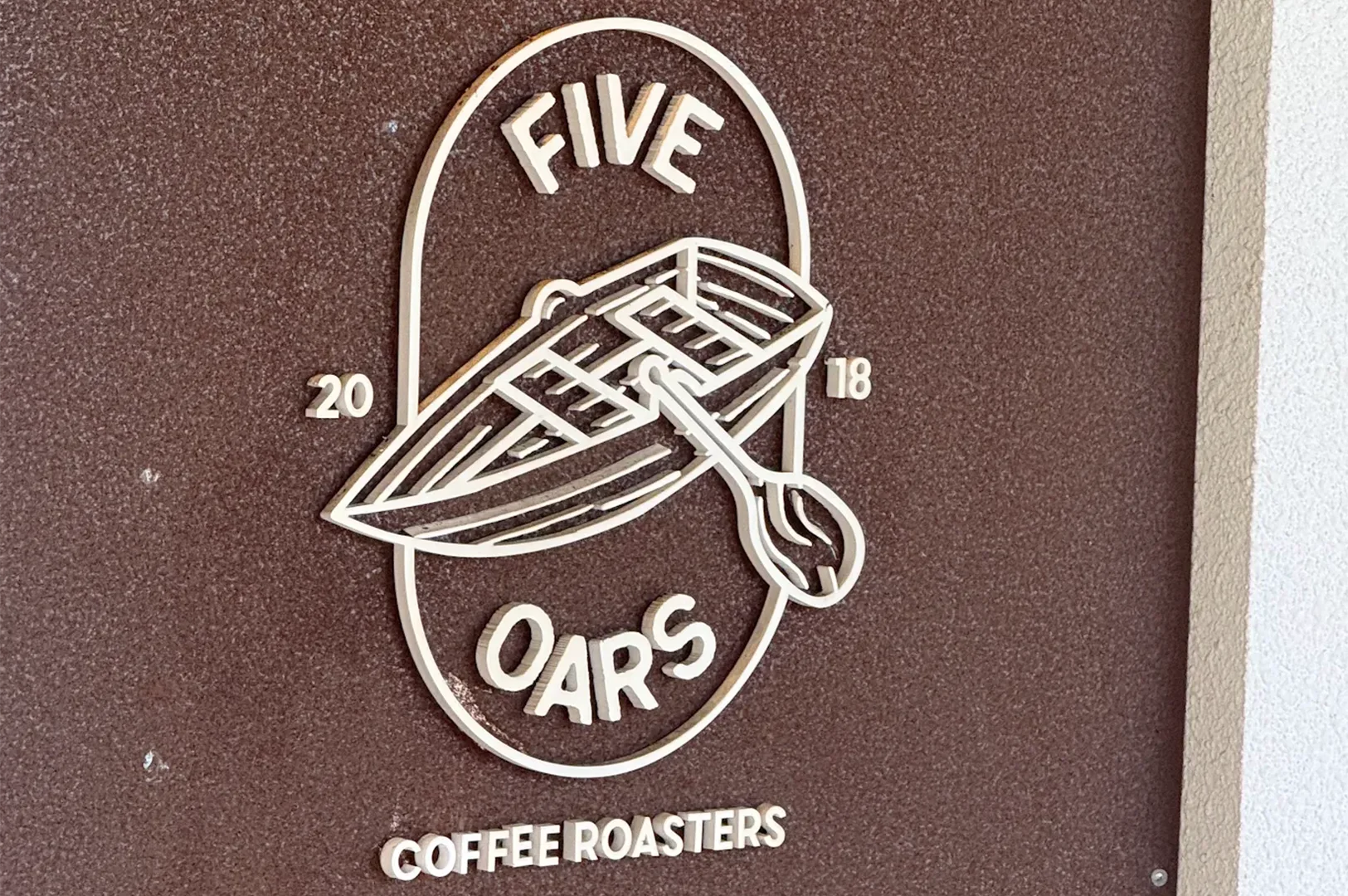 Close-up of the wooden laser-cut logo sign for Five Oars Coffee Roasters established in 2018, mounted on a textured brown wall.