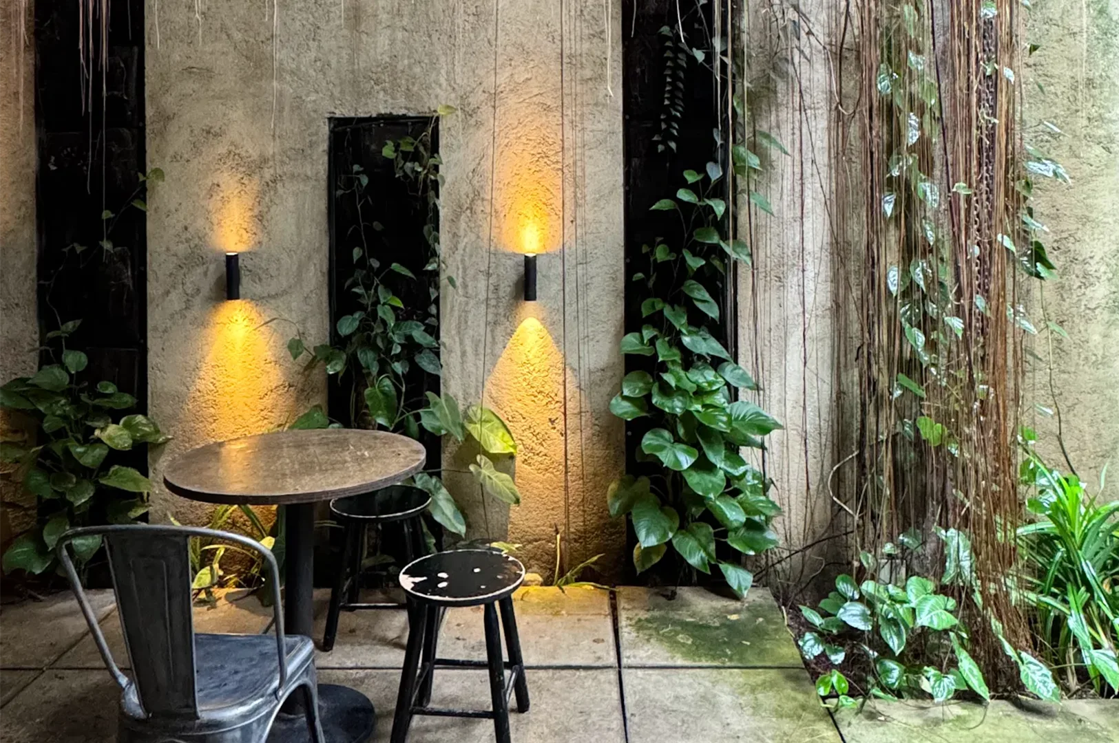 A rustic indoor courtyard cafe seating area with a wooden table, metal chairs, hanging vines, warm wall sconces, and lush green plants.