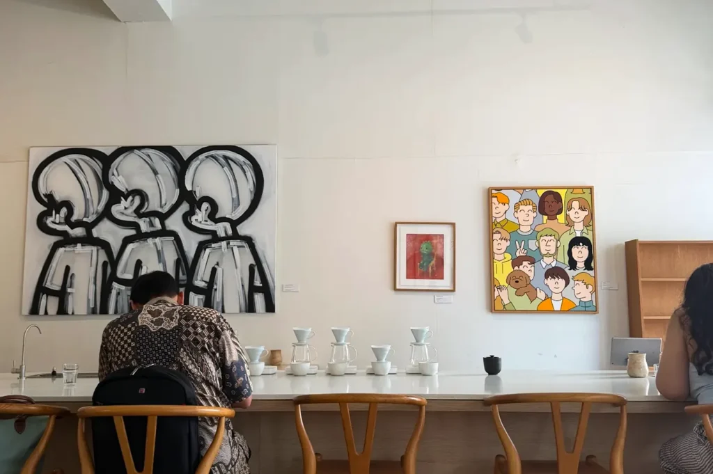 The brew bar counter at Apartment Coffee with customers seated on wooden wishbone chairs, featuring distinctive modern paintings on a white gallery wall and a precise coffee setup.