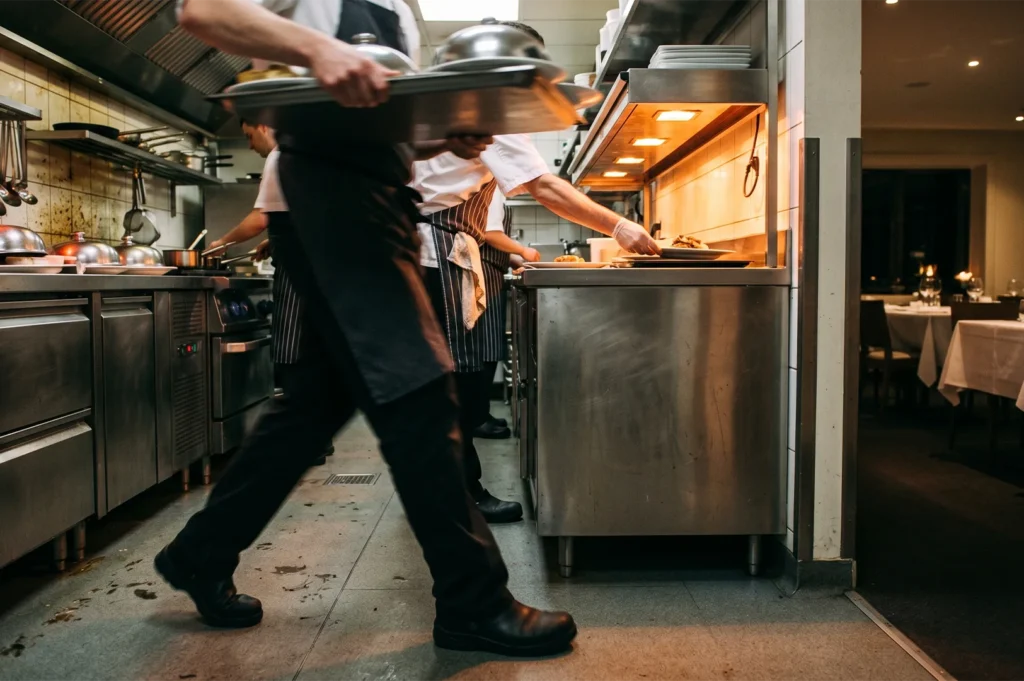 Action shot of professional chefs working in a bustling commercial kitchen, carrying trays, plating gourmet dishes under pass lights, wearing aprons and uniforms during busy dinner service.