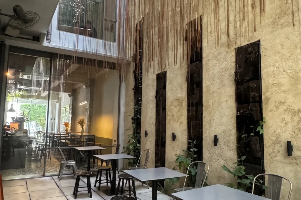 An industrial style cafe interior looking out into a high-ceiling courtyard with metal chairs, dark tables, large glass partitions, and dangling roots.