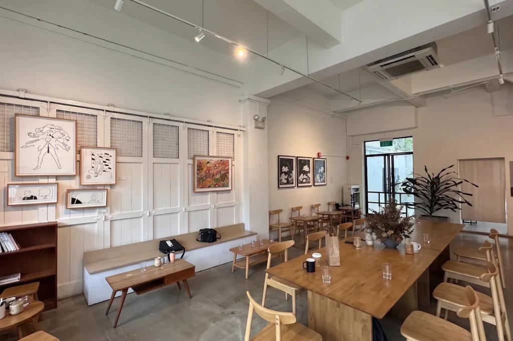 Spacious interior of Apartment Coffee in Singapore highlighting a long wooden communal table, light timber chairs, white paneled walls adorned with art frames, and natural lighting near the exit.