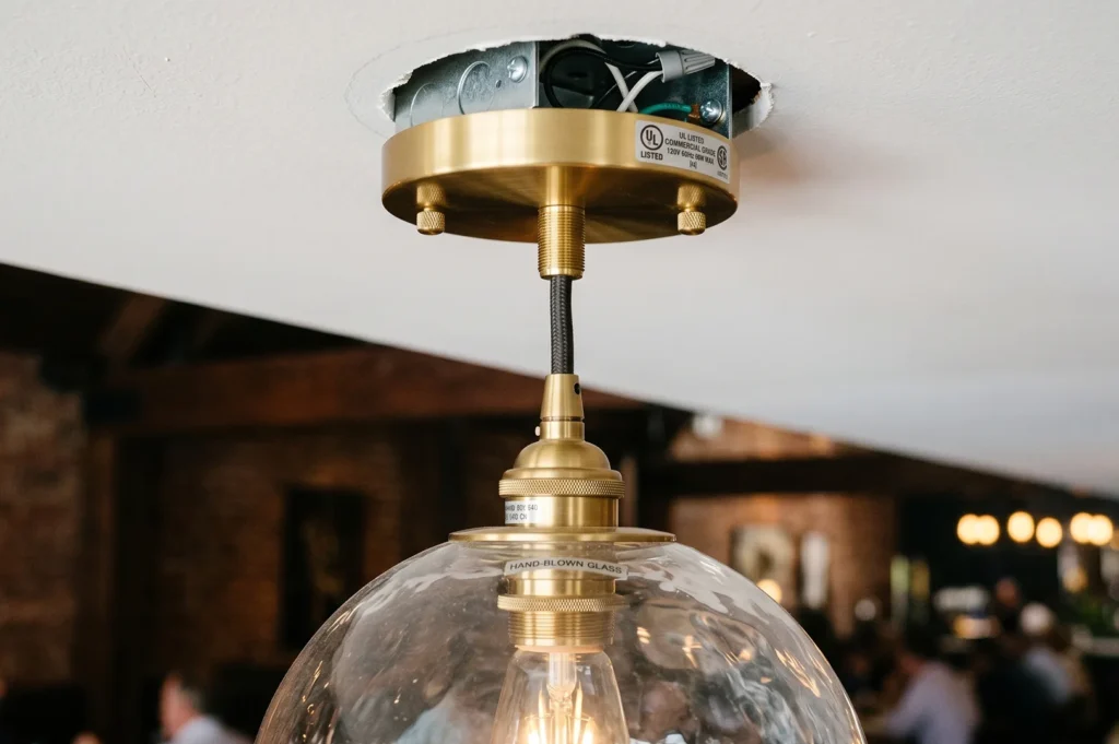 Close-up detail of a brass pendant light fixture installation showing the ceiling electrical box junction, woven cord, and a hand-blown glass globe shade.