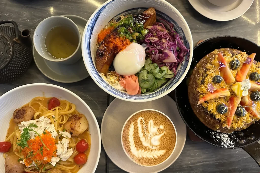 Flat lay view of a rich brunch spread featuring a salmon donburi bowl, scallop and crab pasta, a soufflé pancake, hot tea, and a cafe latte with intricate art.