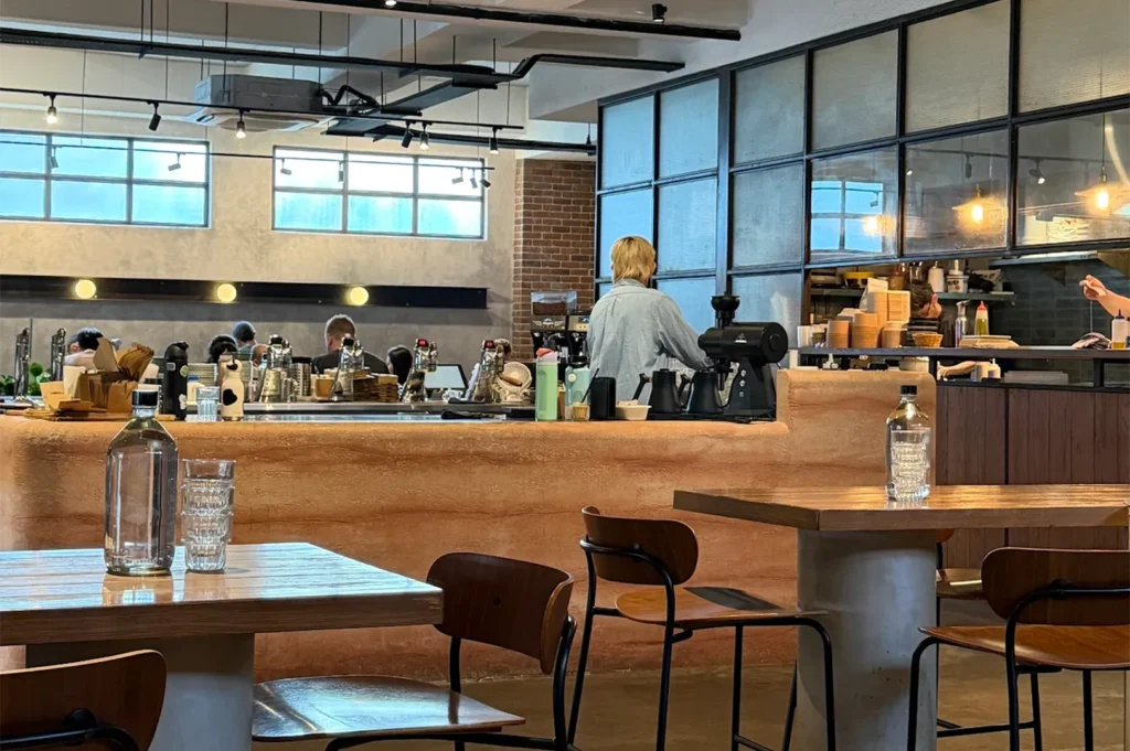 Spacious dining area inside Five Oars Coffee Roasters with wooden tables, industrial lighting, and a long rammed-earth style service counter with staff working.