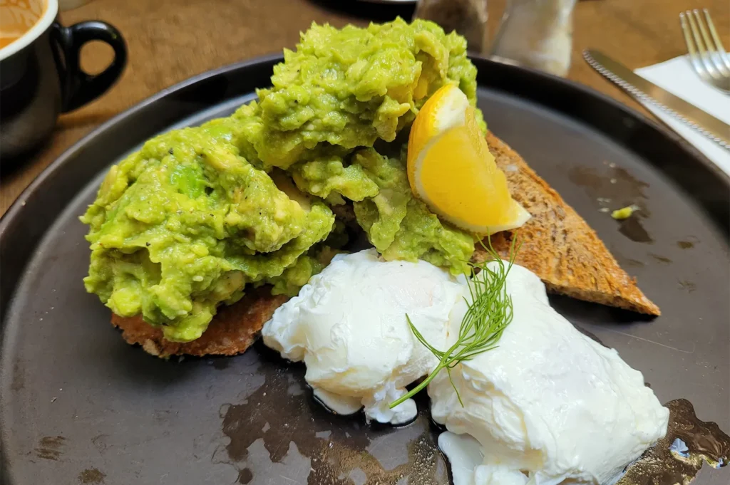A delicious gourmet breakfast plate featuring a generous heap of smashed avocado on toast, two perfectly poached eggs, a fresh dill sprig, and a lemon wedge.
