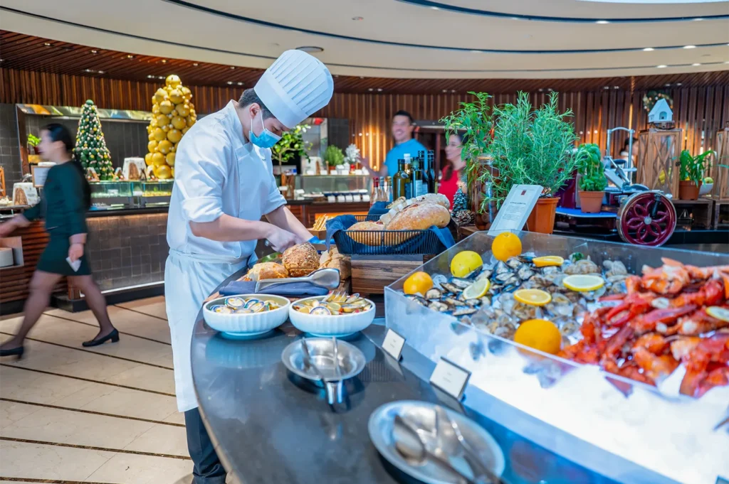A professional hotel chef in uniform carving fresh bread at a luxury buffet station featuring a large ice display of fresh seafood, including lobsters, oysters, and mussels garnished with citrus fruits.