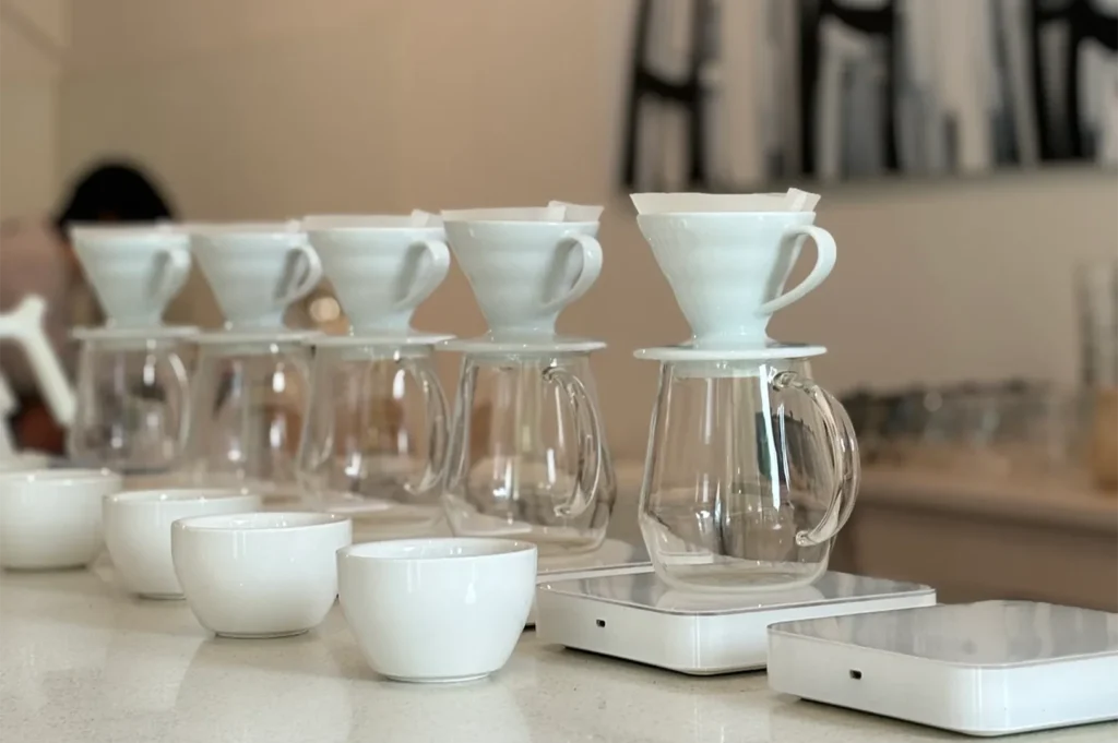 A shallow depth-of-field close-up shot of white ceramic V60 coffee drippers stacked on clear glass servers and white electronic scales at a professional brew bar.