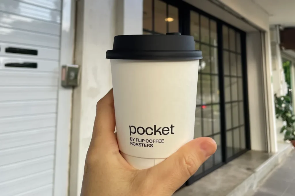 A person holding a white takeaway coffee cup featuring the minimalist black logo of Pocket by Flip Coffee Roasters.