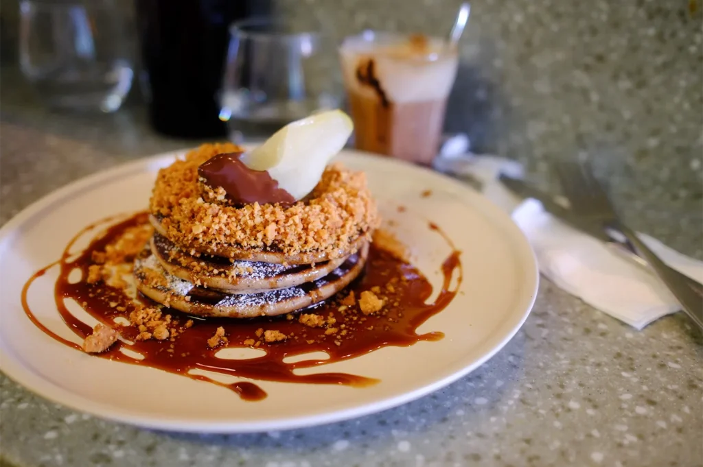 A stack of fluffy pancakes drizzled with rich caramel sauce, topped with crunchy cookie crumble and a chocolate-dipped pear slice on a ceramic plate.