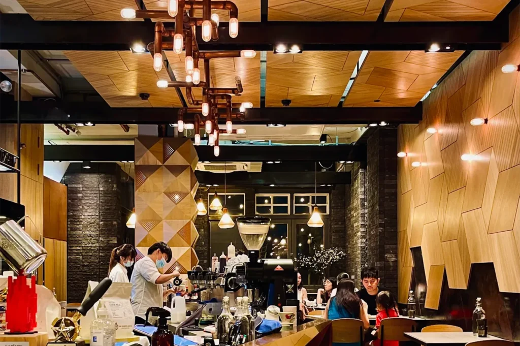 Cozy and bustling interior of a modern specialty coffee shop with industrial copper pipe lighting fixtures and geometric wood paneled walls.
