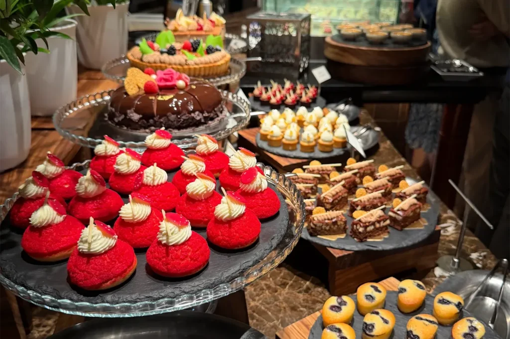 An elegant dessert buffet spread featuring bright red cream-filled choux pastries, chocolate glazed cake, mini fruit tarts, chocolate brownies, and blueberry muffins displayed on premium slate platters.