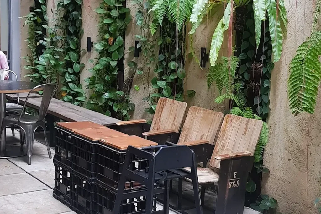 An outdoor cafe patio seating area with wooden theater-style chairs, upcycled milk crate tables, and a vibrant vertical garden with climbing green ivy.