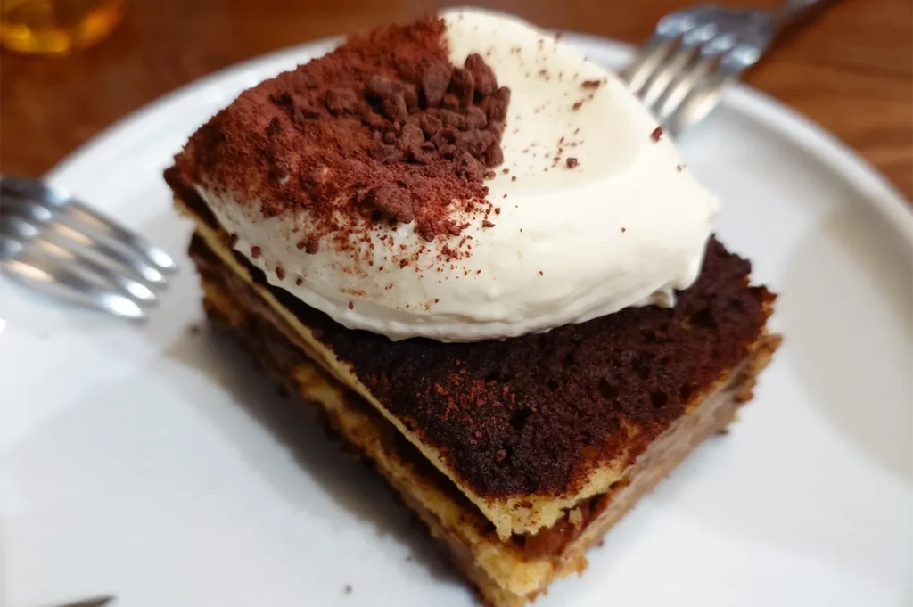 A close-up shot of a rich slice of tiramisu cake topped with generous fresh cream and cocoa powder on a white plate.