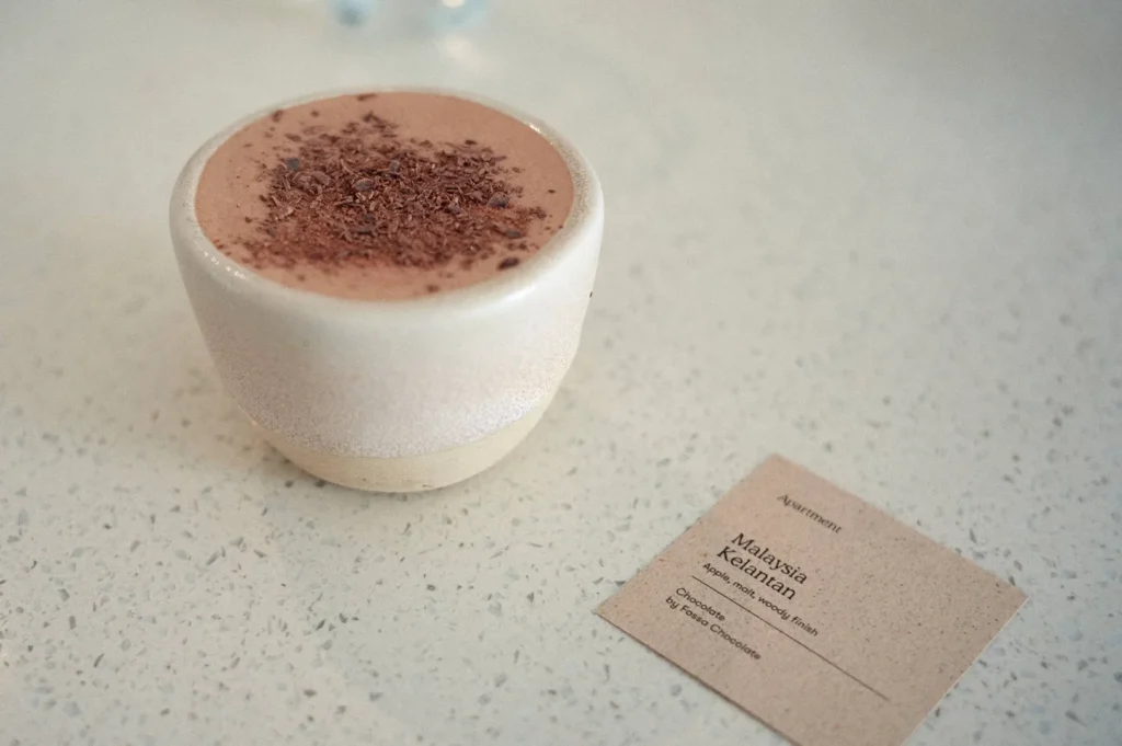 A artisanal cup of hot chocolate dusted with fine shavings served alongside a descriptive origin card for Malaysia Kelantan chocolate by Fossa Chocolate at Apartment Coffee.