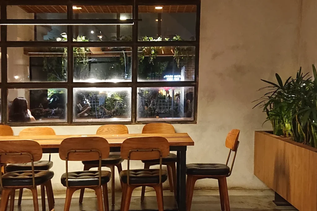 Cozy warm night view of a long wooden dining table and chairs set against a large grid window overlooking the street at Five Oars Coffee Roasters.