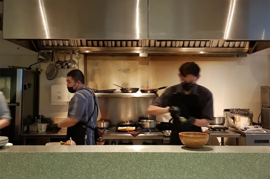 Professional chefs wearing masks and aprons preparing gourmet dishes in a clean, commercial stainless steel cafe kitchen.