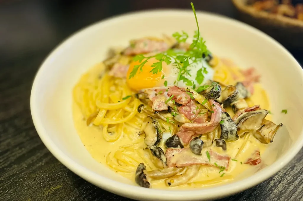 A white bowl filled with creamy carbonara pasta noodles tossed with bacon strips, wild mushrooms, fresh herbs, and topped with a soft poached egg yolk.