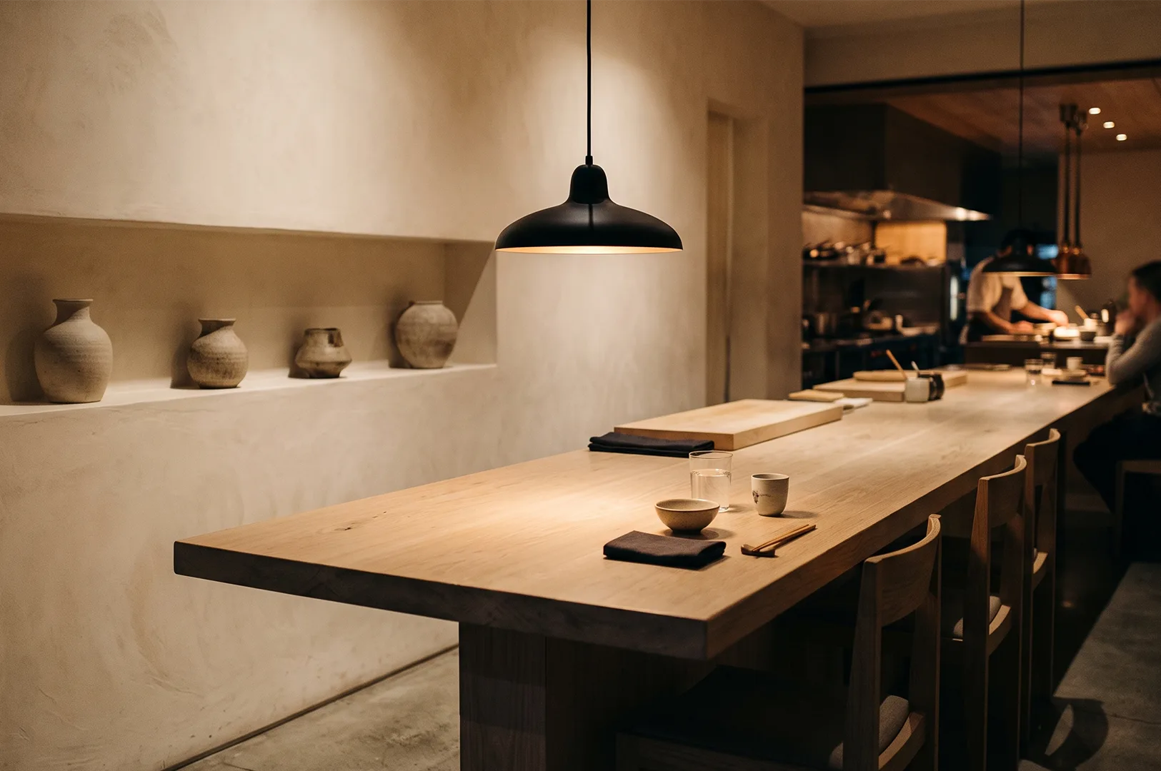 Minimalist fine dining restaurant interior featuring a long light oak chef's table counter under a black pendant light with ceramic vases on a textured wall shelf and kitchen in the background.